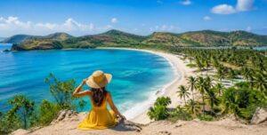 Woman in yellow dress sitting on a hill overlooking a stunning white sand beach and turquoise sea in Lombok dry season