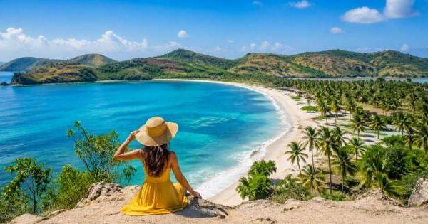 Woman in yellow dress sitting on a hill overlooking a stunning white sand beach and turquoise sea in Lombok dry season