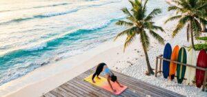 surf and yoga reatreats in kuta lombok. Person practicing a downward dog yoga pose on a wooden deck overlooking Kuta Lombok beach, with ocean waves, palm trees, and surfboards beside them in the morning light.