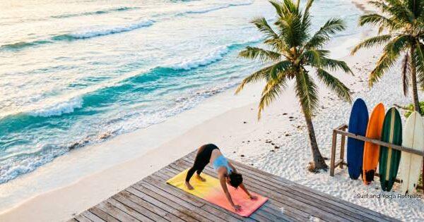 surf and yoga reatreats in kuta lombok. Person practicing a downward dog yoga pose on a wooden deck overlooking Kuta Lombok beach, with ocean waves, palm trees, and surfboards beside them in the morning light.