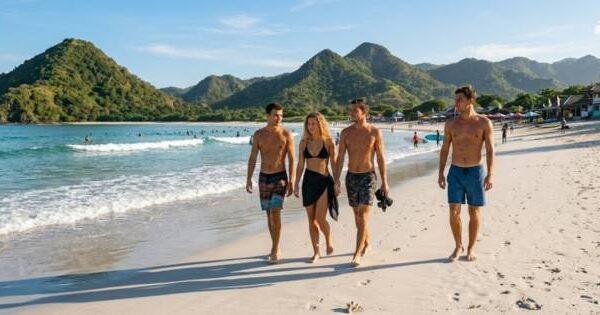 Selong Belanak beach group of travelers walking along white sand shoreline with surfers in water and iconic green pyramid hills in background