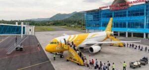 A realistic aerial photograph of Lombok International Airport. A yellow-and-white Scoot aircraft is parked at the terminal with green mountains and a cloudy sky in the background. Passengers are boarding and disembarking the plane. singapore to lombok by scoot